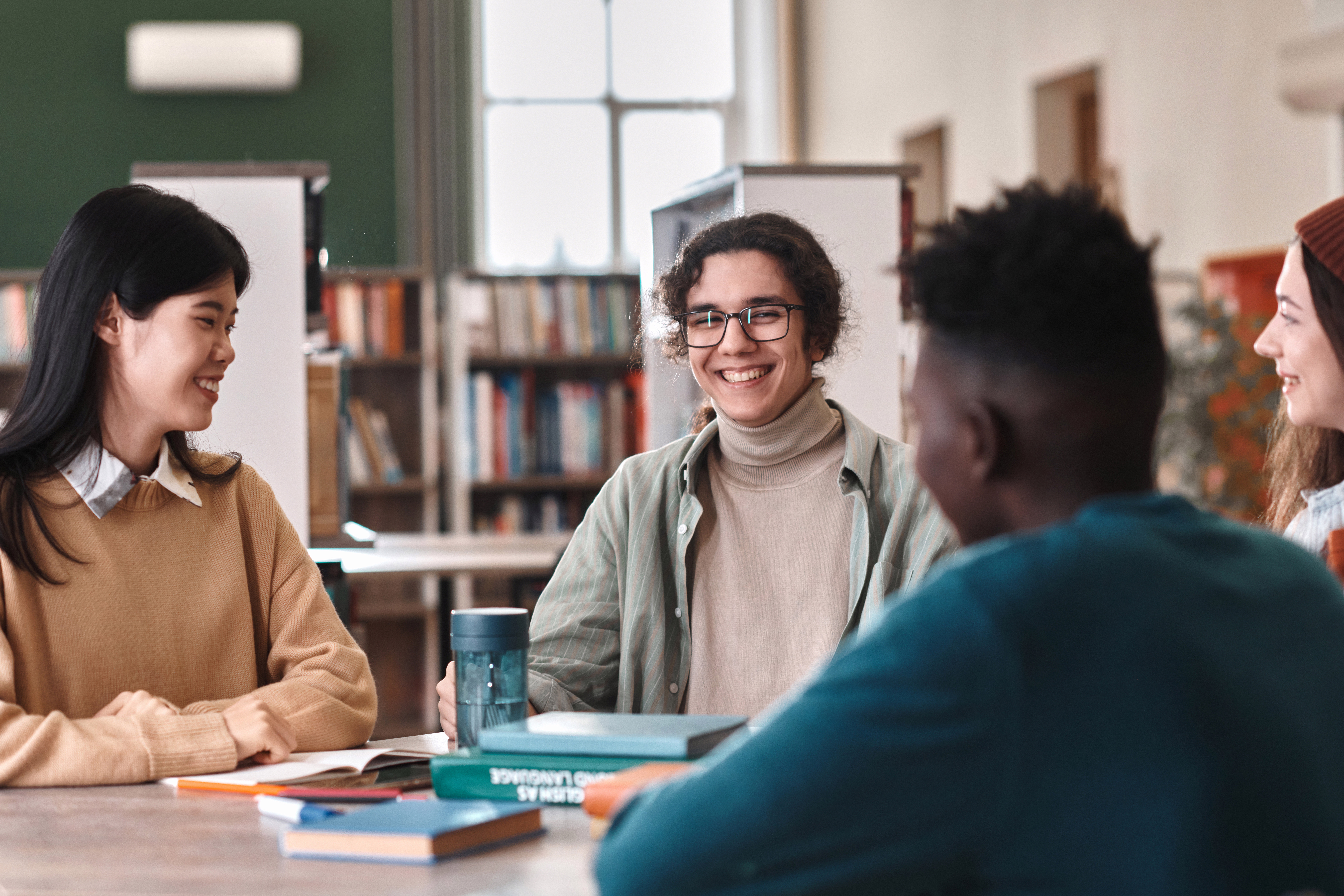 Portrait of young male student with genuine smile enjoying schoolwork sitting at table with friends in library
