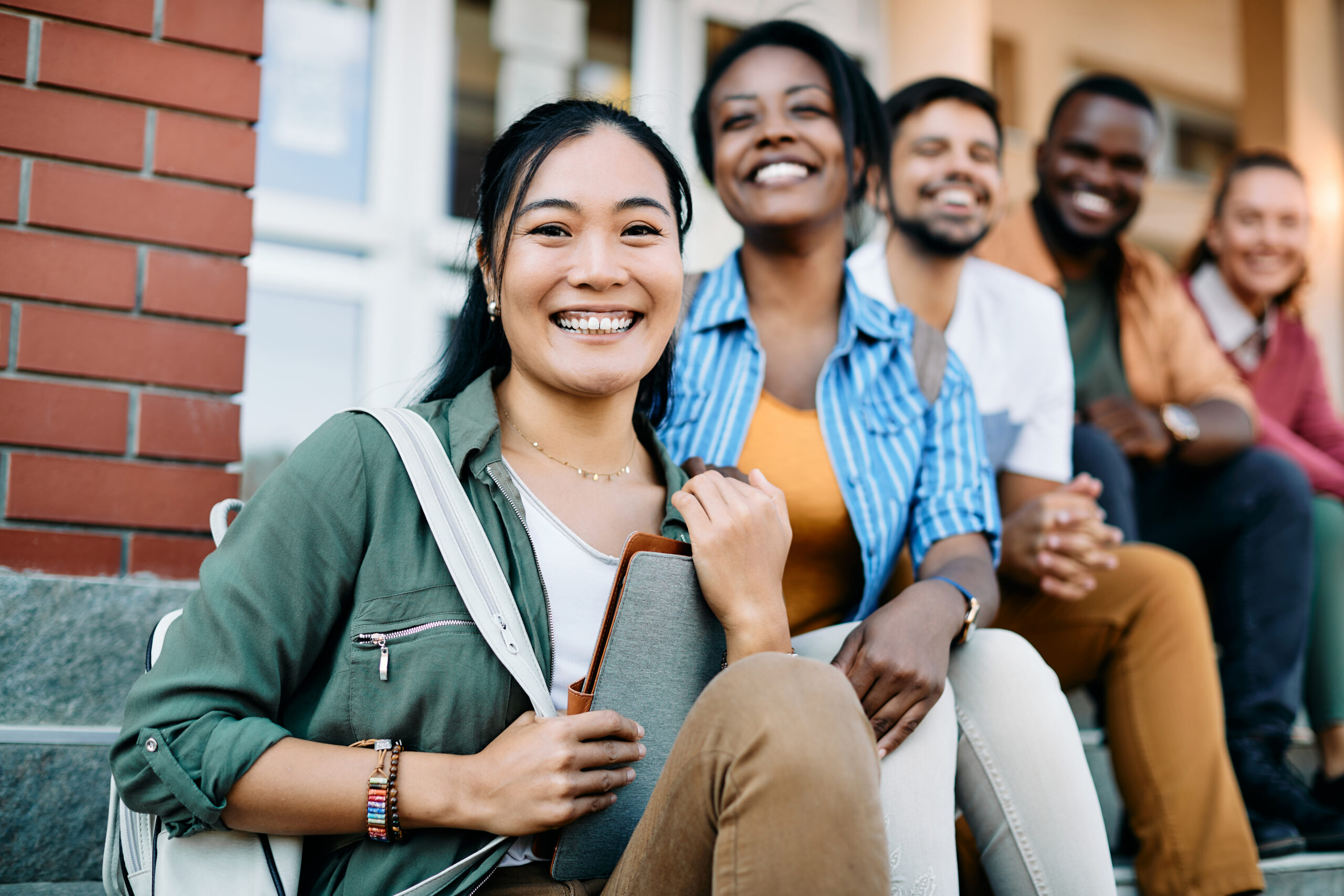 Happy Asian student and her college friends sitting outdoors at campus and looking at camera.